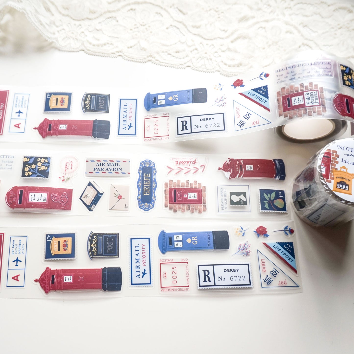 A roll of decorative PET tape featuring vintage-style red and blue British postboxes, letters, and postage stamps, laid out on a white surface with a lace cloth in the background.