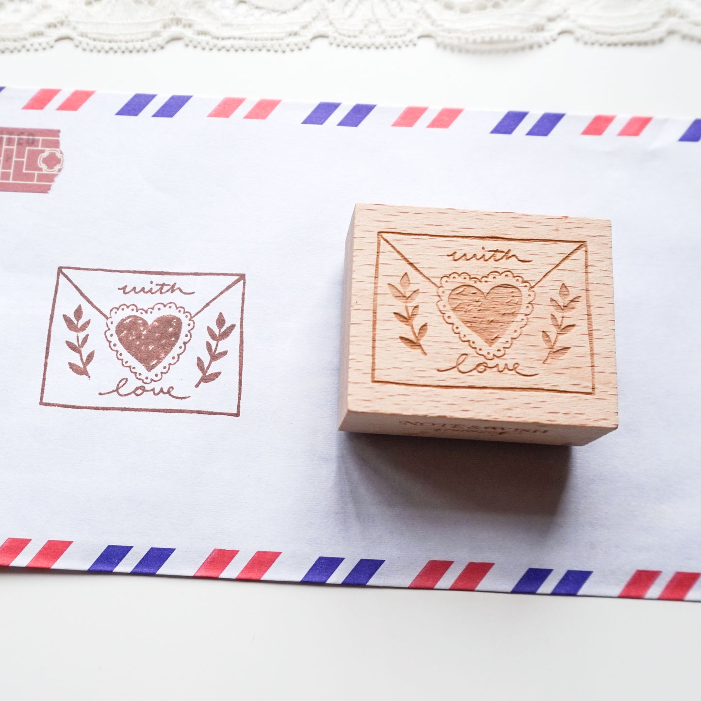 A wooden rubber stamp with an engraved heart-in-envelope design sits next to its printed impression on white paper with red and blue striped borders.