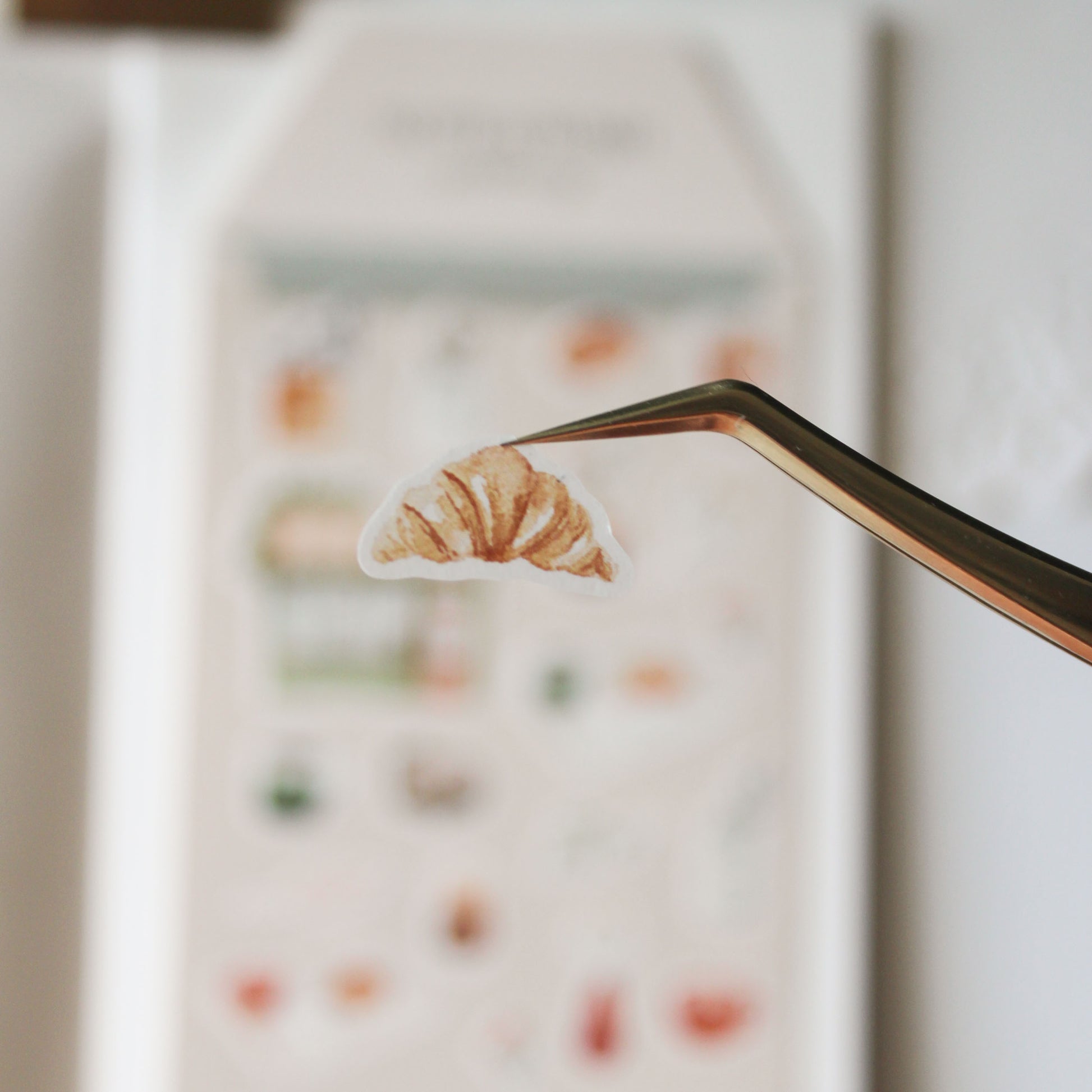 Croissant sticker held by a pair of tongs with a blurred background