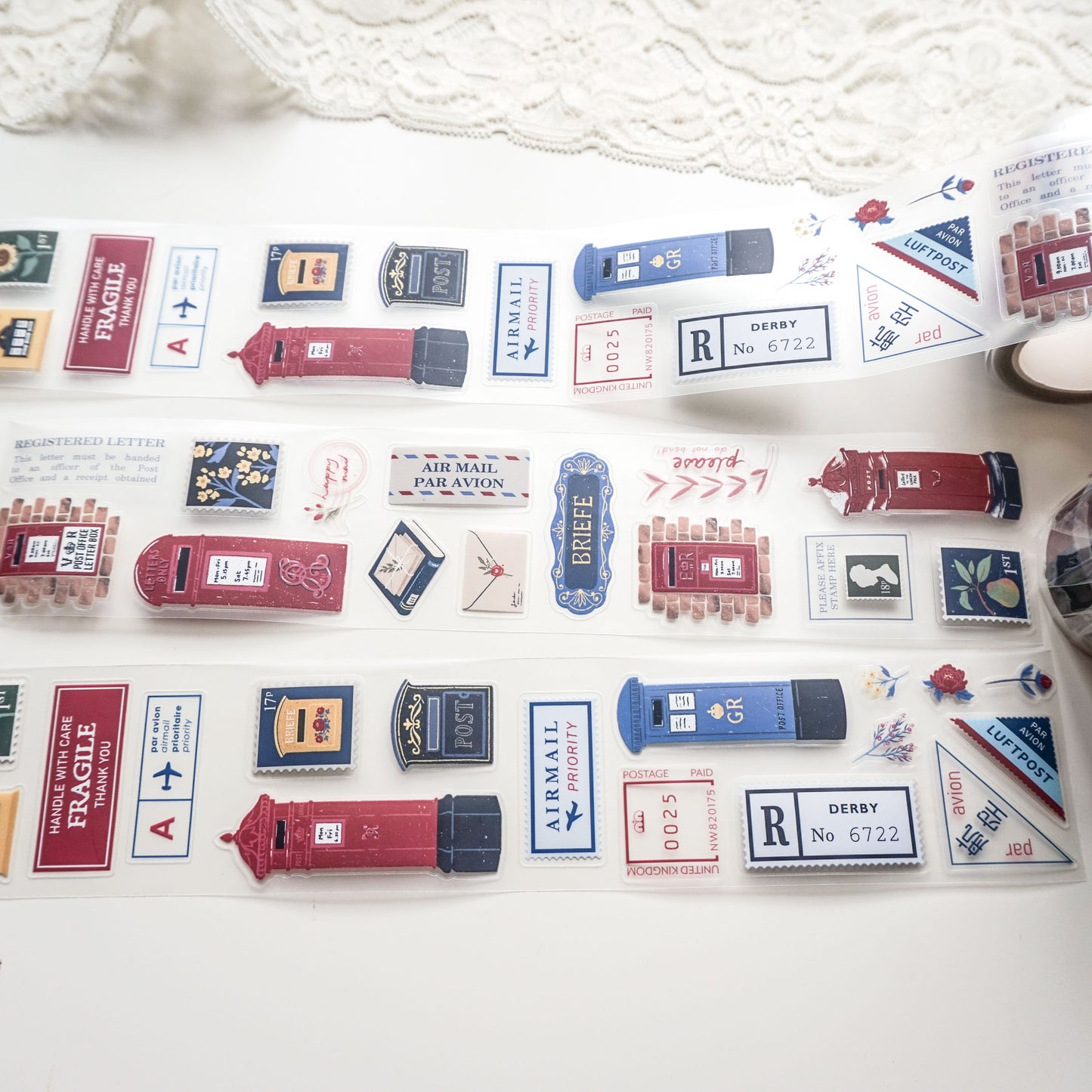 A roll of decorative PET tape featuring vintage-style red and blue British postboxes, letters, and postage stamps, laid out on a white surface with a lace cloth in the background.