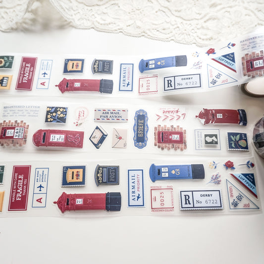 A roll of decorative PET tape featuring vintage-style red and blue British postboxes, letters, and postage stamps, laid out on a white surface with a lace cloth in the background.