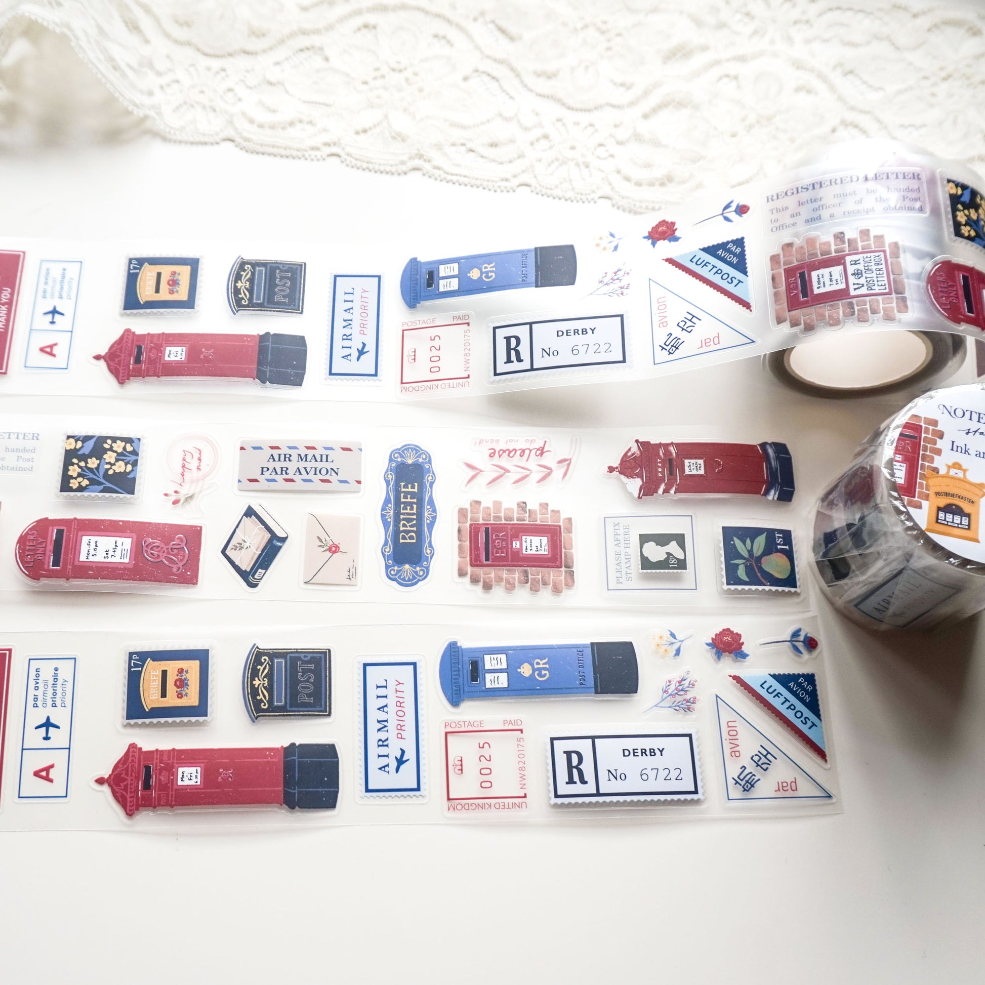 A roll of decorative PET tape featuring vintage-style red and blue British postboxes, letters, and postage stamps, laid out on a white surface with a lace cloth in the background.