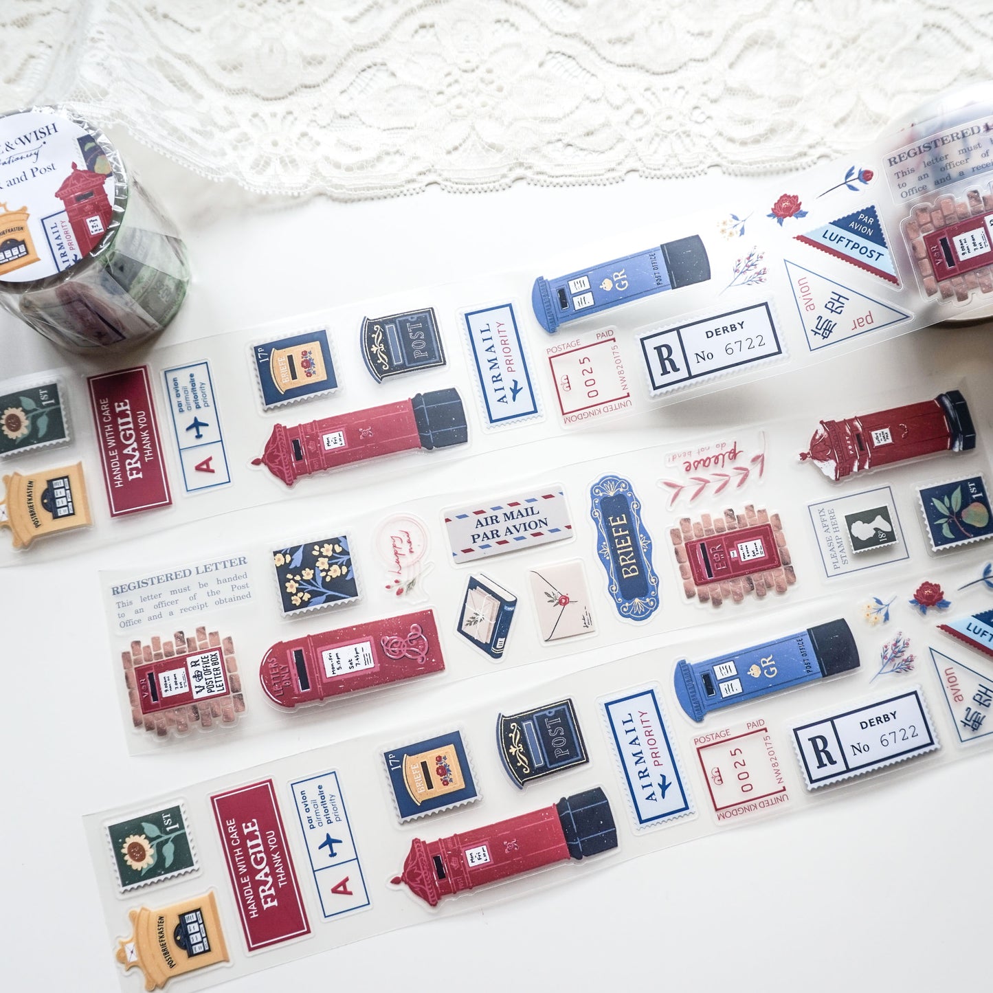 A roll of decorative PET tape featuring vintage-style red and blue British postboxes, letters, and postage stamps, laid out on a white surface with a lace cloth in the background.