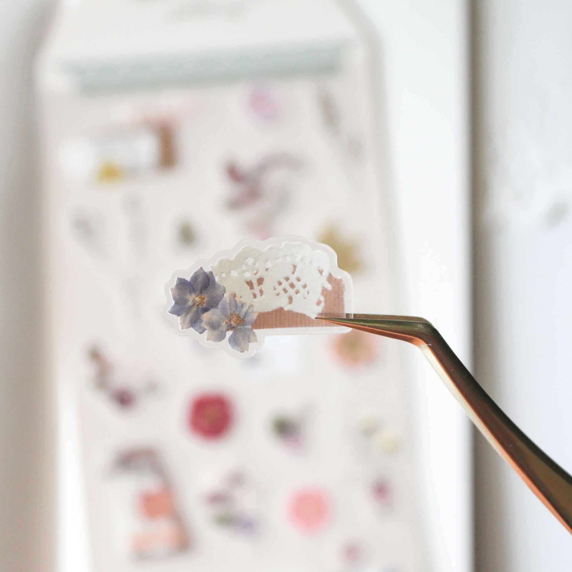Small white lace doily with dried flowers sticker held by tweezers against a blurred background