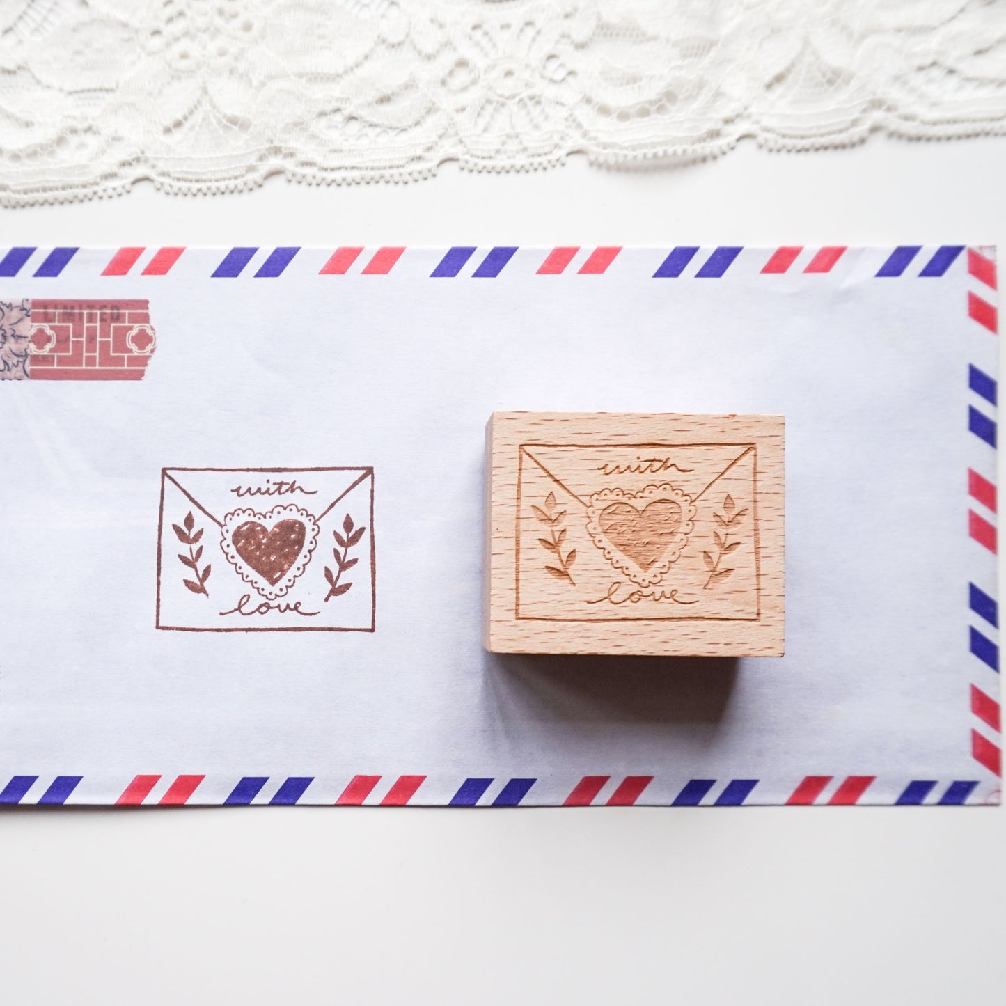 A wooden rubber stamp and its corresponding printed impression on white paper, styled with a red and blue striped airmail envelope and lace trim in the background.