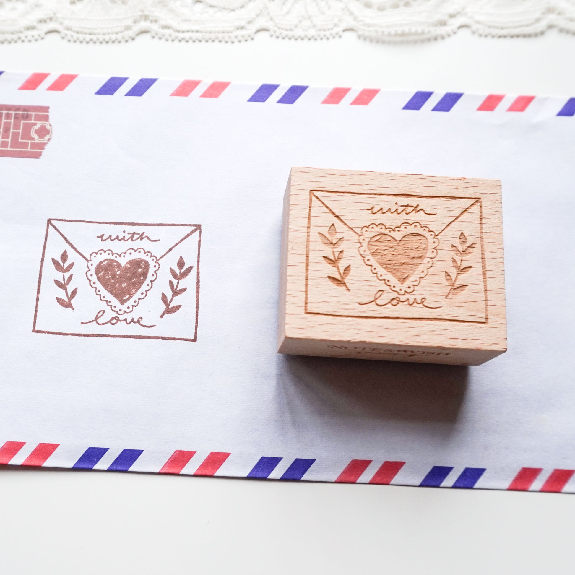 A wooden rubber stamp with an engraved heart-in-envelope design sits next to its printed impression on white paper with red and blue striped borders.
